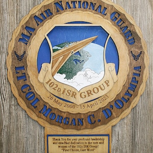 May include: A wooden plaque with a blue and brown design. The plaque features a blue shield with a white jet plane flying over a green and white landscape. The text "MA AIR NATIONAL GUARD 102d ISR GROUP 20 May 2000 - 15 April 2023 LT COL MORGAN C. D'OLYMPIA" is inscribed on the plaque.
