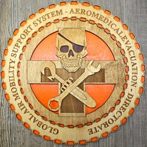 May include: A wooden round sign with an orange background and a brown wooden skull with an eye patch. The skull is crossed with two wrenches. The sign has the text "GLOBAL AIR MOBILITY SUPPORT SYSTEM - AEROMEDICAL EVACUATION - DIRECTORATE" around the outside.