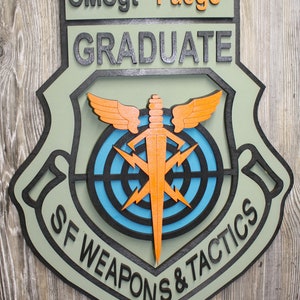 May include: A green, black, and orange wooden plaque with the text "CMSgt "Fuego" GRADUATE SF WEAPONS & TACTICS" and a stylized image of a target with lightning bolts and a dagger.