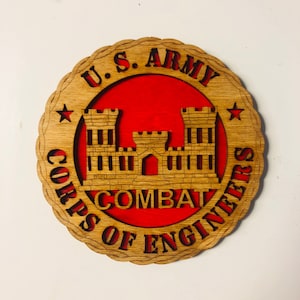 May include: A wooden round sign with a red background and the words "U.S. ARMY CORPS OF ENGINEERS" and "COMBAT" in a brown font. The sign features a stylized image of a castle with two towers.