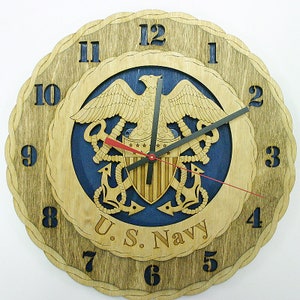 Puede incluir: Reloj de pared de madera con el emblema de la Marina de los EE. UU., que presenta un águila, un escudo y anclas. El reloj tiene un diseño en capas con un centro azul y un anillo exterior marrón claro. Los números negros marcan las horas.