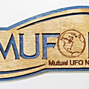 Puede incluir: Un letrero de madera con un borde azul y el texto "MUFON Mutual UFO Network" en una fuente estilizada. El letrero presenta un pequeño globo terráqueo en el centro.