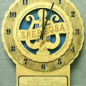 Puede incluir: Un reloj de madera con un diseño azul y marrón. La esfera del reloj presenta el texto "Society for the Preservation and Encouragement of Barber Shop Quartet Singing in America" y el acrónimo "S.P.E.B.S.Q.S.A.". El reloj también presenta una placa con el texto "WC Walter Thank You for your service Above and Beyond the call of Duty to the A Capella Fellas 2018".