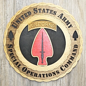 May include: Wooden plaque featuring the words "UNITED STATES ARMY SPECIAL OPERATIONS COMMAND" in a circular design. The center displays a red arrowhead with a black dagger, and the word "AIRBORNE" is above it.