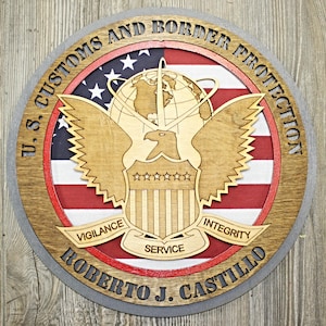 May include: A round wooden plaque with a gray border, featuring a red, white, and blue American flag design. The center of the plaque displays a stylized eagle with a globe and the text "U.S. Customs and Border Protection". Below the eagle are the words "Vigilance", "Service", and "Integrity". The bottom of the plaque reads "Roberto J. Castillo".
