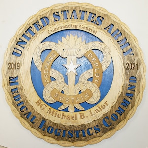 Może przedstawiać: Drewniana tabliczka z brązowym tłem i niebieskim tekstem. Tabliczka przedstawia logo Dowództwa Logistyki Medycznej Armii Stanów Zjednoczonych, z tekstem "Commanding General" i "BG Michael B. Lalor". Zawiera również lata 2019 i 2021.