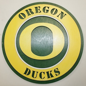 May include: A circular wooden sign with a yellow outer ring and a green inner ring. The inner ring features a white outline of the letter "O" with a green center. The words "OREGON" and "DUCKS" are written in white letters on the sign.