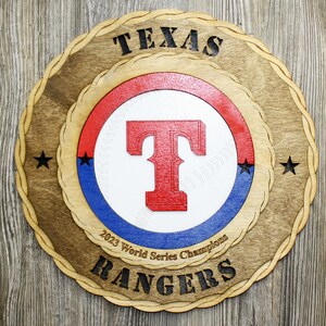 May include: A wooden plaque with a baseball-themed design. It features a red, white, and blue circle with a white baseball and a red "T" in the center. The text "TEXAS RANGERS" is written in black around the circle. The text "2023 World Series Champions" is written in black below the circle.