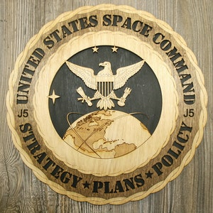 May include: A wooden plaque with a circular design featuring the United States Space Command logo. The logo includes a bald eagle with wings spread, a globe, and the text "United States Space Command J5 Strategy * Plans * Policy J5".