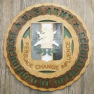 May include: A wooden round sign with a brown background and green accents. The sign features the text "U.S. ARMY PSYCHOLOGICAL OPERATIONS" in a circular pattern around the edge. In the center is a shield with a white horse and the text "PERSUADE CHANGE INFLUENCE".