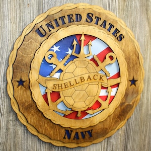 May include: Wooden United States Navy emblem with a layered design. The emblem features the words "United States" and "Navy" in blue, surrounding a central design with a turtle, anchors, keys, and the word "Shellback". The background includes the American flag.