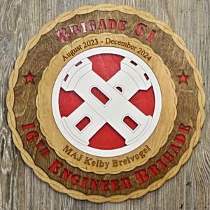May include: A wooden plaque with a red and white emblem of two crossed towers. The plaque is inscribed with the text "16th Engineer Brigade", "MAJ Kelby Breivogel", "Brigade SI", "August 2023 - December 2024".