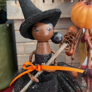 May include: A black witch doll with a black hat and a black and orange dress holding a broom. The doll has brown skin and black eyes.