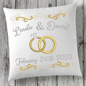 以下が含まれることがあります： 「Linda & David February 24th 2023」という刺繍が施された白い枕。結婚指輪とハートも刺繍されています。