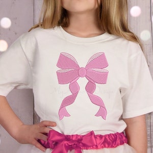 May include: A white t-shirt with a pink embroidered bow on the front. The bow is large and has a center knot.