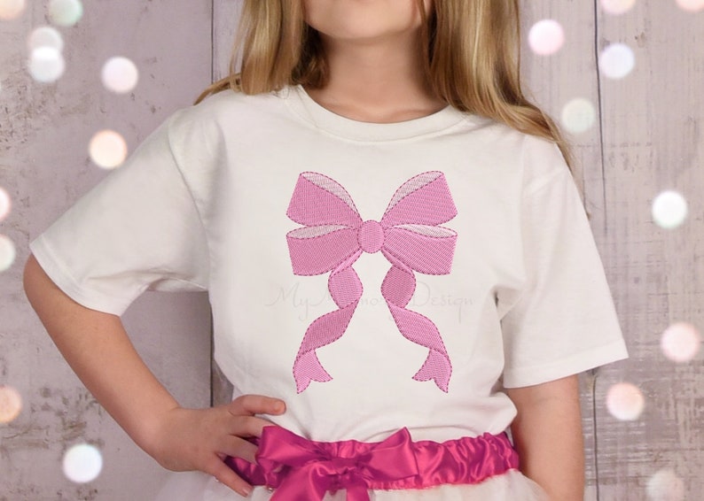 May include: A white t-shirt with a pink embroidered bow on the front. The bow is large and has a center knot.