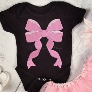 May include: A black baby onesie with a pink embroidered bow on the front. The onesie is laying on a white wooden surface with a pink tulle skirt and a pink flower.