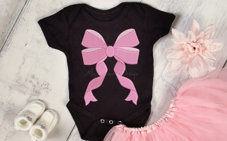 May include: A black baby onesie with a pink embroidered bow on the front. The onesie is laying on a white wooden surface with a pink tulle skirt and a pink flower.