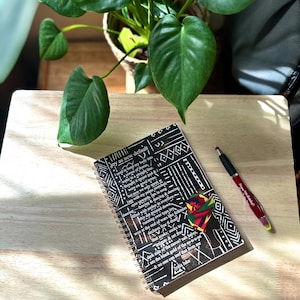 May include: A black and white spiral notebook with a colorful design on the cover. The notebook has a quote about joy and dancing on the cover. The notebook is sitting on a wooden table with a green plant and a red pen.