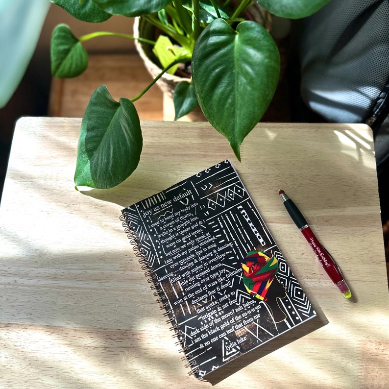 May include: A black and white spiral notebook with a colorful design on the cover. The notebook has a quote about joy and dancing on the cover. The notebook is sitting on a wooden table with a green plant and a red pen.