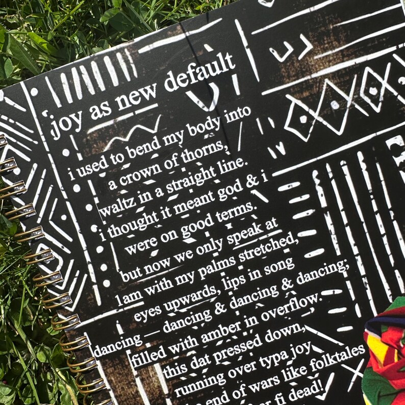 May include: A black and white spiral bound journal with a black background and white geometric patterns. The journal has the text "joy as new default" printed on the front cover. The text is in white and is centered on the cover. The journal is open to a page with black and white text.