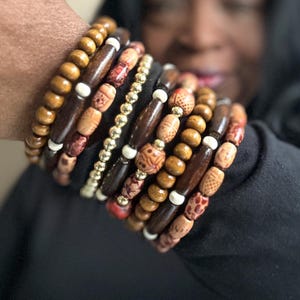 May include: A collection of wooden beaded bracelets in shades of brown, tan, and red. Some beads are smooth, while others have carved patterns. A gold beaded bracelet is also included. The bracelets are worn on a person's arm.