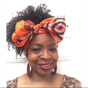 May include: A woman with curly black hair is wearing a large orange and brown patterned head wrap tied in a bow. She is wearing large red earrings.