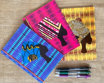 May include: Three notebooks with colorful patterned covers featuring a silhouette of a woman's head with a patterned head wrap. The notebooks are stacked on top of each other and there are three pens next to them.