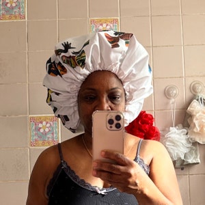 May include: A white satin shower cap with a colorful print of stylized figures. The cap is worn by a person holding a gold smartphone.