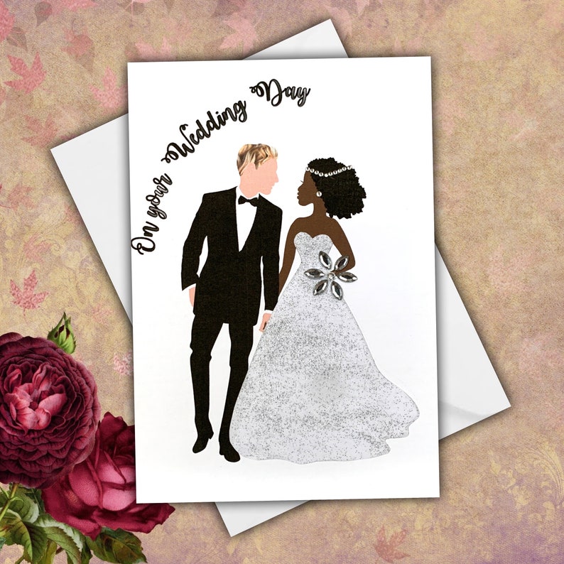 A wedding card with an interracial couple. The white groom is wearing a black tuxedo, he has  blonde hair. The black bride is wearing a white glitter a-line dress, She has curly shoulder length hair. The text on the card reads 'On your Wedding Day'.