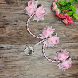 May include: A pink and silver beaded necklace with a silver charm in the shape of a crown and a Mickey Mouse head. The necklace is attached to two pink and silver hair bows with clips.