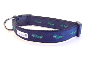 May include: A navy blue dog collar with a repeating pattern of green alligators. The collar has a silver D-ring and a black plastic buckle. The collar is labeled "Pawsitivewear".