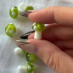 Puede incluir: Una colección de pequeñas setas de vidrio verde con lunares blancos. Las setas miden aproximadamente 2,5 cm de alto y tienen una tapa redonda y un tallo blanco.