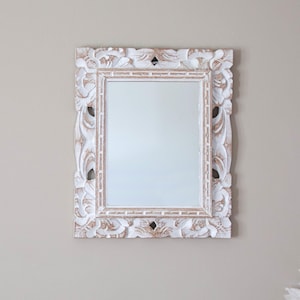 May include: White wooden mirror with an ornate carved frame. The mirror has a rectangular shape and is hanging on a wall.