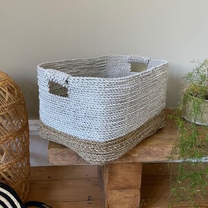 May include: A rectangular woven basket with a two-tone design. The top portion is a light gray, and the bottom is a natural brown. The basket has built-in handles and is sitting on a wooden surface.