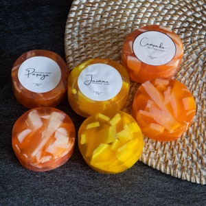 May include: Six round natural soap bars with different scents and fruit pieces embedded in the soap. The soaps are arranged on a woven wicker mat. The scents are Papaya, Jasmine, and Cempaka.