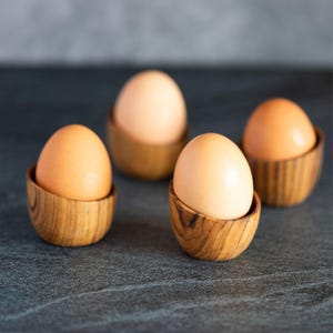 May include: Four brown eggs nestled in small, wooden egg cups. The eggs are light brown, and the cups are made of light-coloured wood with a visible grain. The arrangement is on a dark, textured surface.