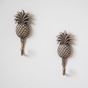 May include: Two bronze pineapple-shaped wall hooks. Each hook has a detailed pineapple design with a textured surface and a hook at the bottom. The hooks are mounted on a white wall, offering a tropical decor element.