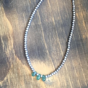 May include: A silver-toned necklace with small, round beads and a series of teardrop-shaped green gemstones. The necklace is displayed on a wooden surface, showcasing its delicate design and color contrast.