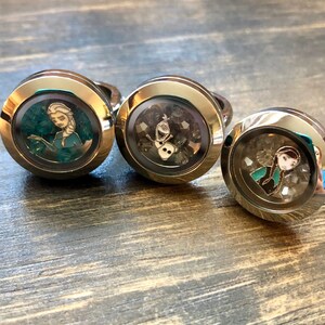 May include: Three silver locket rings with images of characters from the movie Frozen. The first ring features Elsa, the second ring features Olaf, and the third ring features Anna.