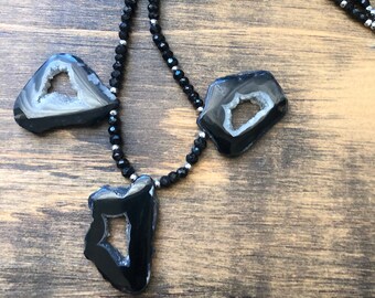 May include: A black beaded necklace with three agate pendant charms. The charms are black with a white agate center and a hole in the center. The necklace is on a wooden surface.