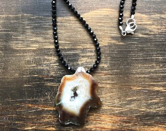 May include: A black beaded necklace with a brown and white agate pendant. The pendant has a natural, irregular shape with a hole in the center.