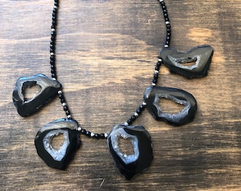 May include: A black necklace with a silver clasp and five black agate slices with white crystal inclusions. The agate slices are irregularly shaped and have a rough texture.