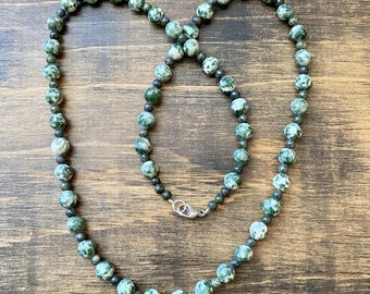 May include: A green and white stone beaded necklace with a silver clasp. The necklace is made of round beads and is on a wooden surface.