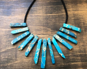 May include: A turquoise and white patterned stone necklace with a black bead chain. The necklace features a fan-shaped design with multiple stone pieces.