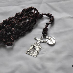 Może przedstawiać: Brązowy różaniec ze srebrnym krucyfiksem i medalikiem Świętego. Koraliki różańca są wiązane, a krucyfiks przedstawia Jezusa. Medalik przedstawia wizerunek świętego i napis "Saint".