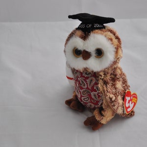 May include: A brown and white owl plush toy wearing a black graduation cap that says "Class of 2023". The owl is wearing a red and white paisley patterned shirt. The toy has a Ty tag attached to its wing.
