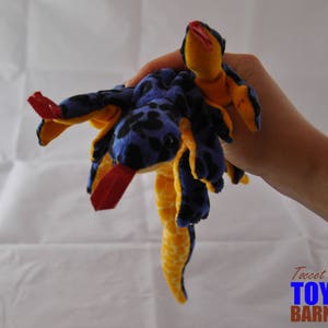 May include: A plush toy snake in shades of blue, yellow, and red. The snake has a blue body with black spots, yellow accents, and a red tongue. The toy is held in a hand.