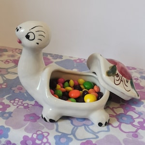 Puede incluir: Un plato de cerámica blanca para dulces con forma de tortuga con un diseño de flor rosa en la tapa. El plato está abierto y lleno de caramelos de colores.