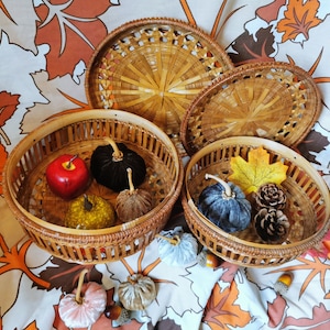 May include: Four woven baskets with lids, each containing a variety of autumn-themed decorations, including small pumpkins, gourds, and a red apple. The baskets are arranged on a patterned fabric background.